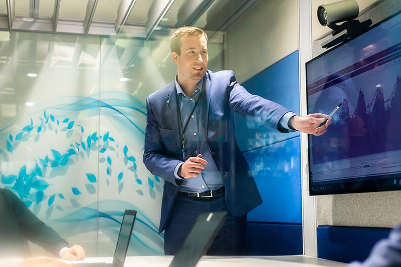 Man wearing a blue suit giving a presentation, pointing at some interesting data on the screen behind him